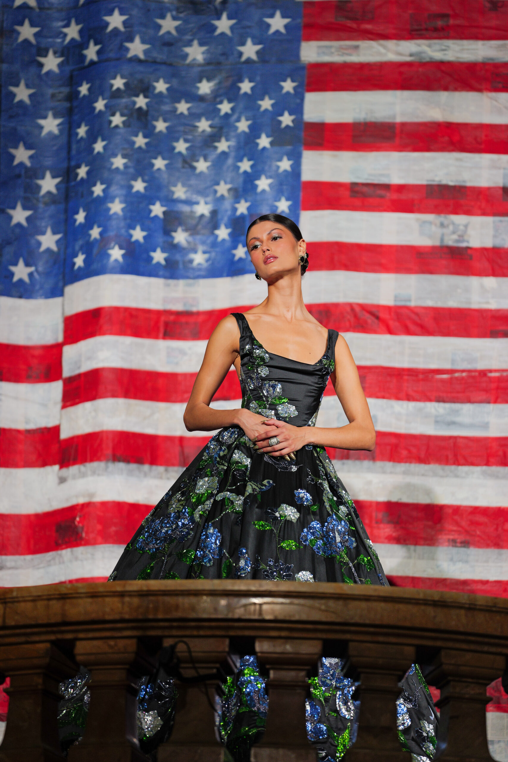Model wearing a black floral gown from Alice + Olivia’s Spring 2026 “The American Woman” collection, standing in front of a large American flag backdrop at Surrogate’s Court during New York Fashion Week.