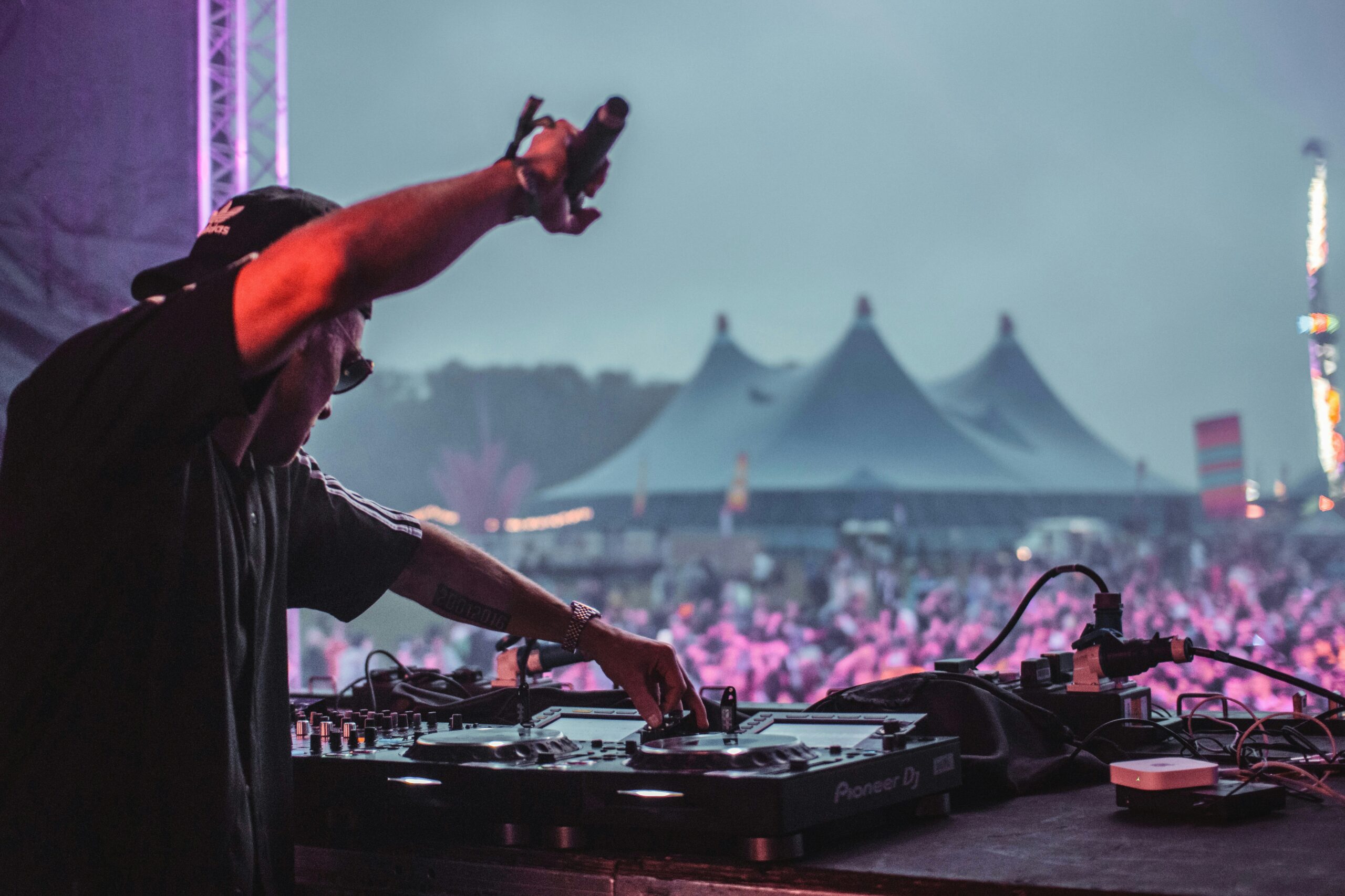 DJ performing on stage at an outdoor music festiva Coachella, operating Pioneer DJ decks with one arm raised toward a massive crowd, festival tents and colourful lights visible in the background at dusk.